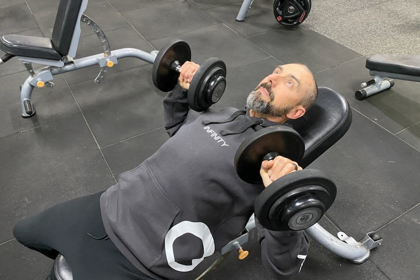 A man doing bench presses with dumb-bells at a gym.