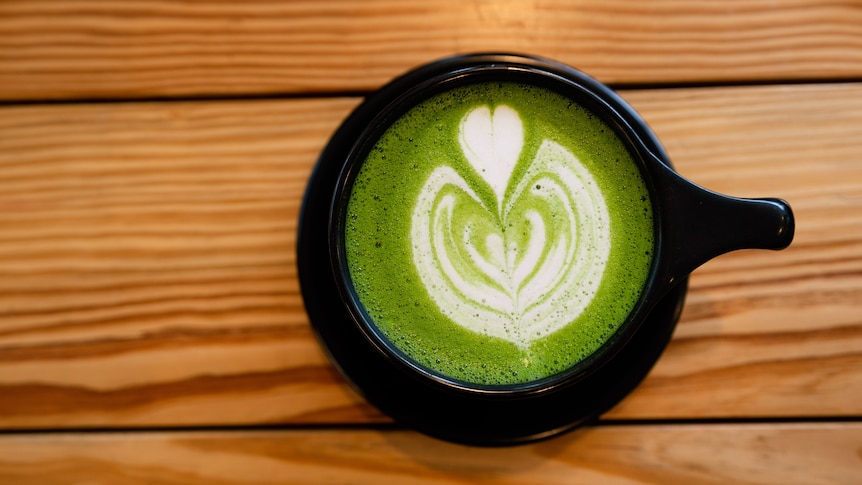 An aerial view of a green-coloured matcha latte in a black cup, sitting on top of a wooden table.