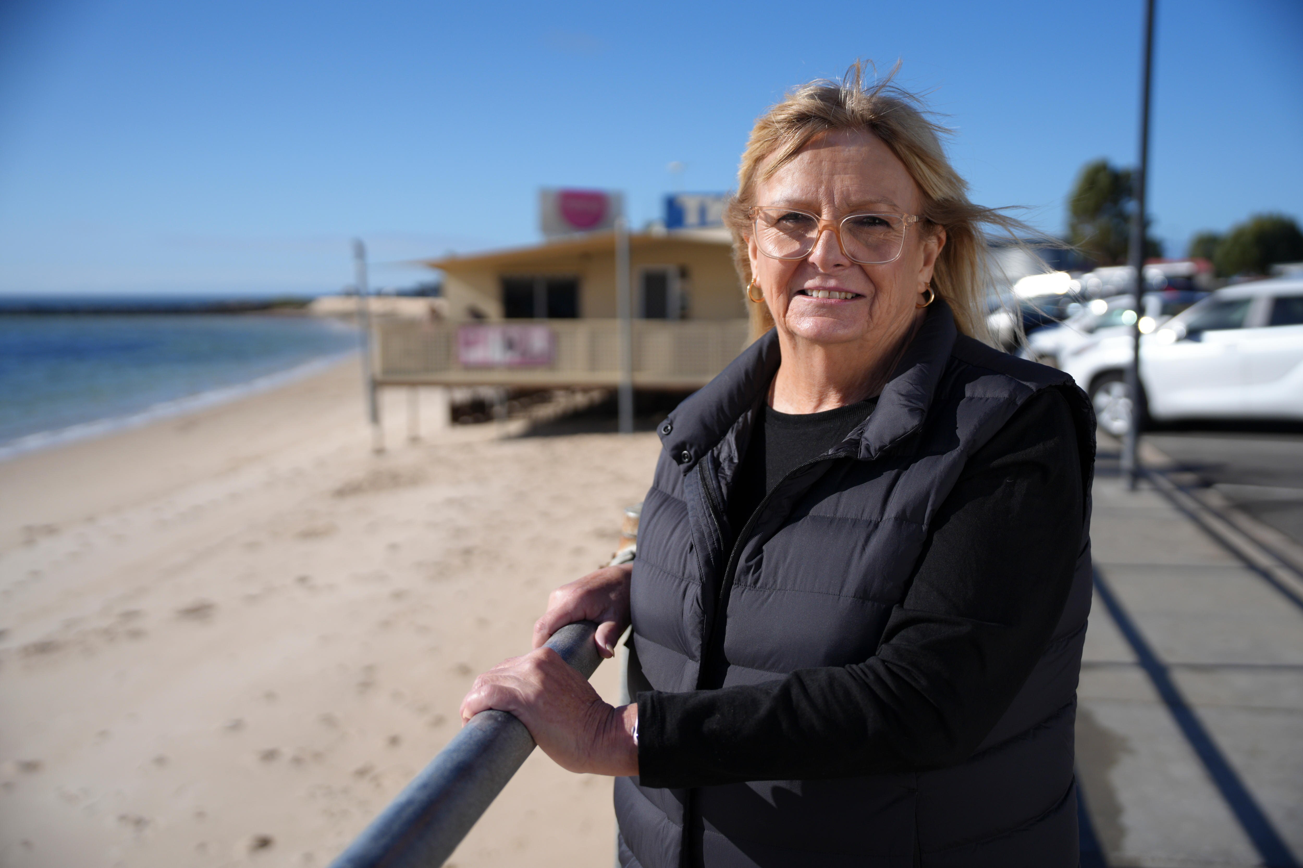 Una mujer sonríe, tiene el cabello rubio soplado por el viento. Ella se apoya en un poste, detrás de ella hay una playa con un quiosco.