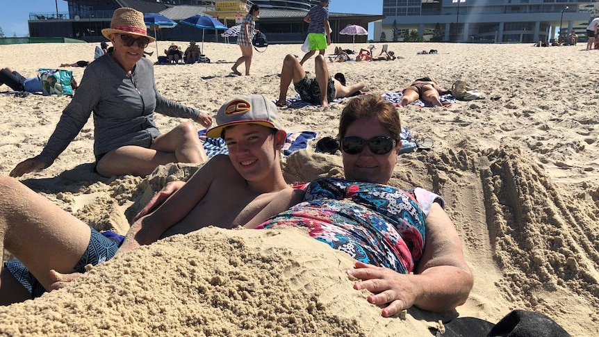 A woman in floral swimwear is buried in sand next to a teenage boy wearing a baseball cap and boardshorts.