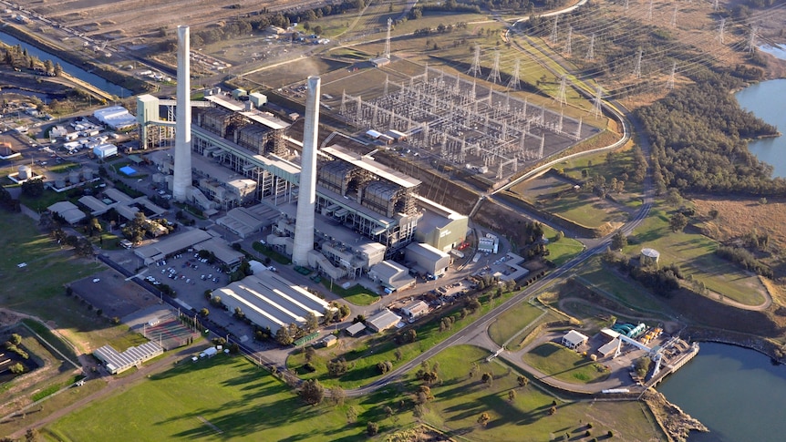 A large power station with two tall concrete smoke stacks surrounded by transmission lines and a lake.