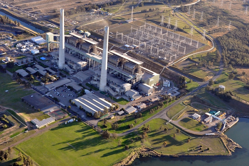 A large power station with two tall concrete smoke stacks surrounded by transmission lines and a lake.