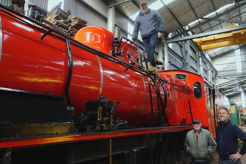 One man atop a loco's boiler and two men on the shed floor below, all smiling to camera.