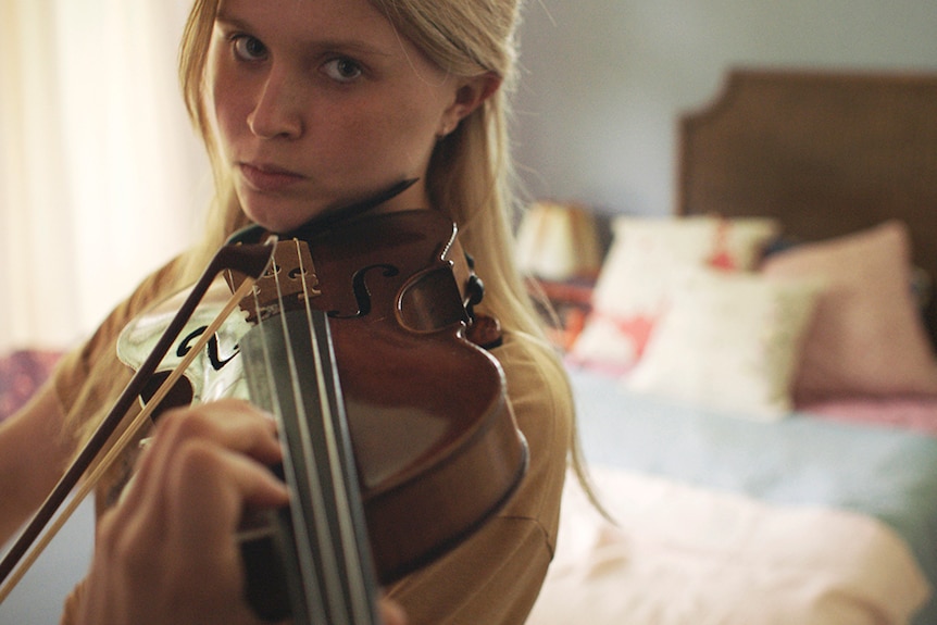 A young blonde woman stands with serious expression in bedroom with violin held between shoulder and chin in playing position.