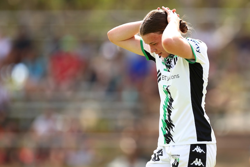 A soccer player wearing white, black and green holds her hands on her head and looks at the ground during a game