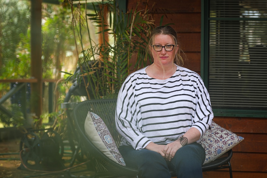 Am MCU of a middle-aged woman with spectacles and a striped shirt posing for a photo in front of plants.