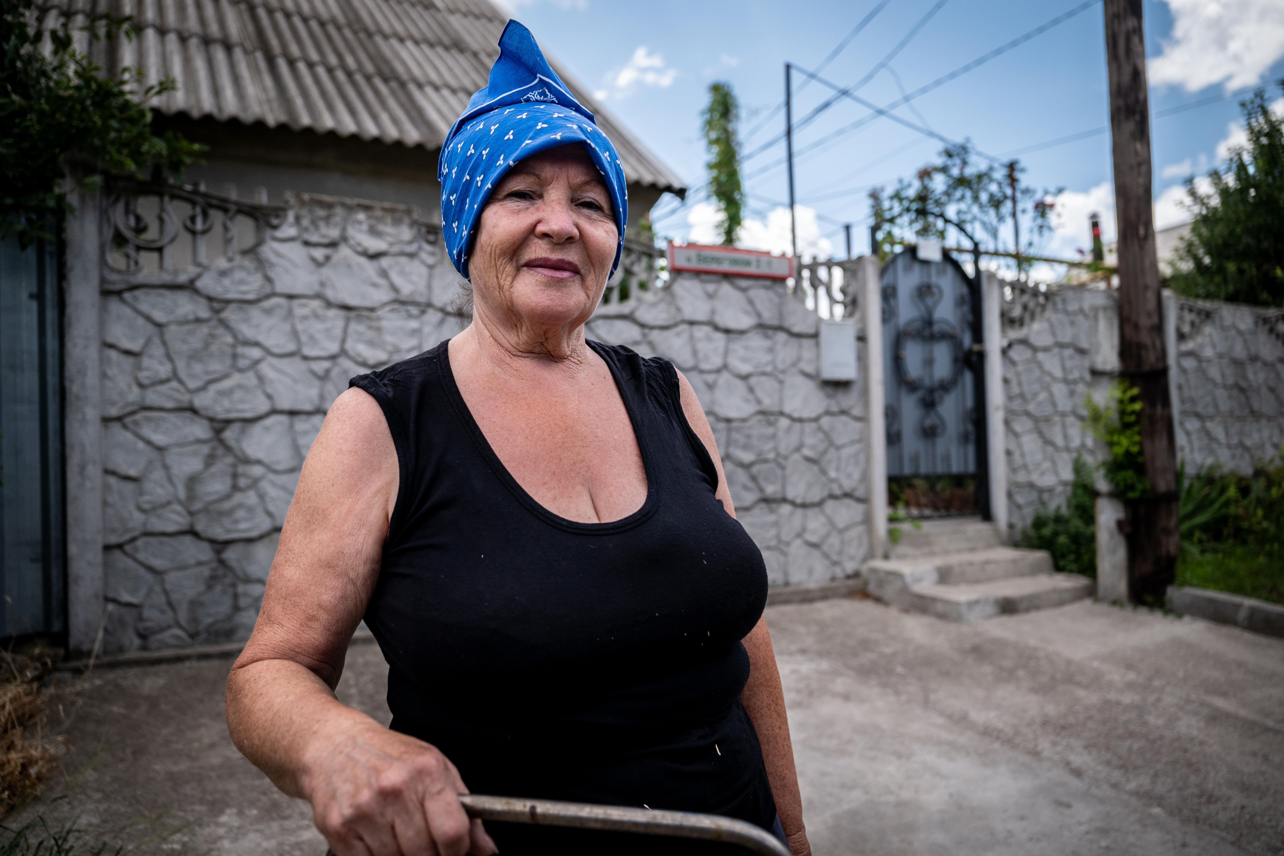 A middle-aged woman wearing a black tank top and blue bandana stands outside a brick wall with ornate gate