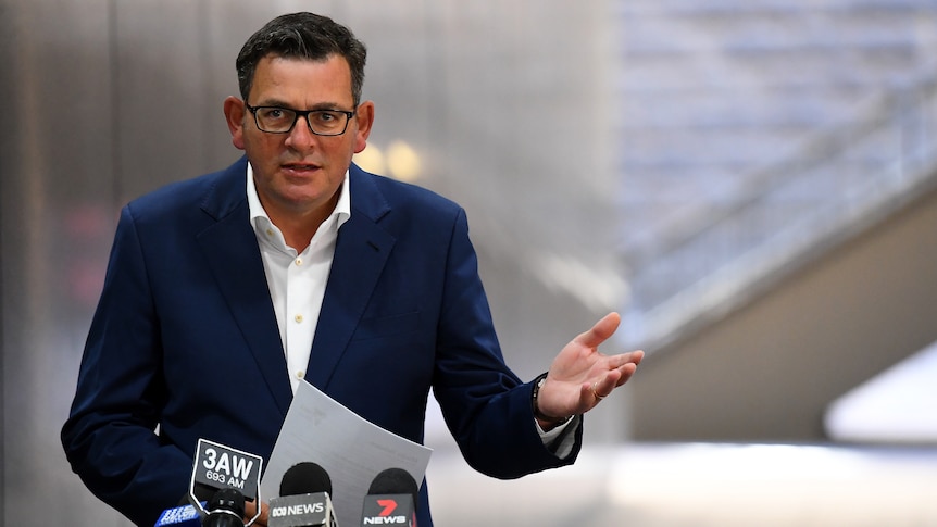 Daniel Andrews, dressed in a white shirt and blue jacket, speaks in front of a microphone stand outside.