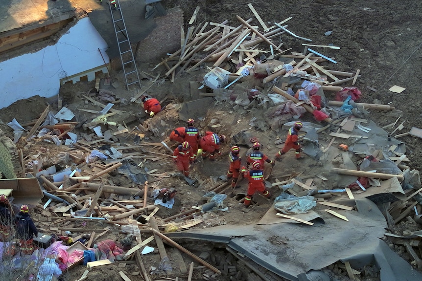 An aerial view of people in red emergency worker suits in the rubble of a collapsed building.
