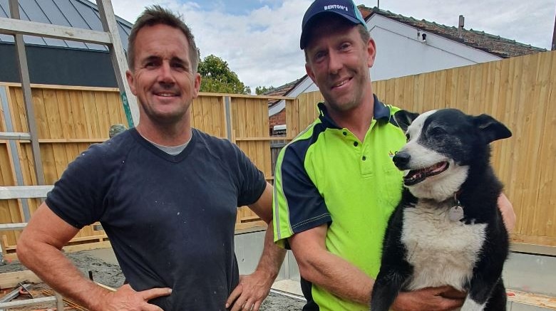 A picture of two tradesmen on a work site with concentre in the background. One man holds a dog.