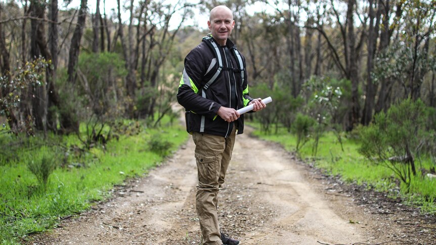 A bald man in brown pants and a black jacket standing on a country road, holding a roll of paper.