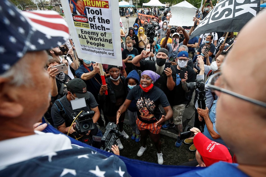 A crowd of people wearing masks holding placards condemning Trump looks up at two people.