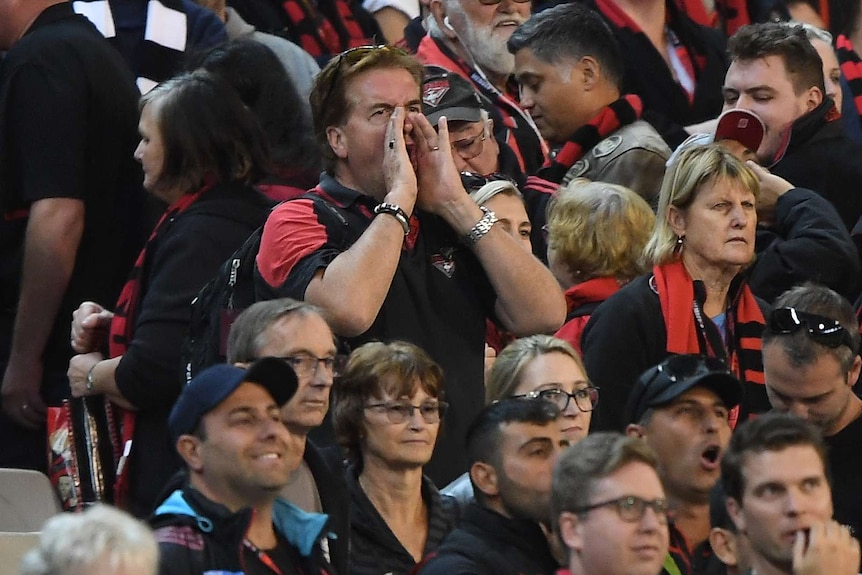 Supporters wearing black and red shirts and scarves cup their hands around their mouths and boo