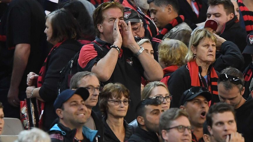 Supporters wearing black and red shirts and scarves cup their hands around their mouths and boo