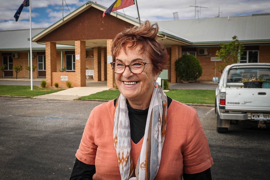 A woman stands in front of a hospital wearing a coral top, red hair and glasses. 