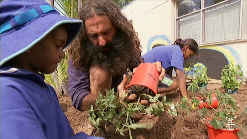 Man showing young boy in hat how to plant a pot plant in the garden