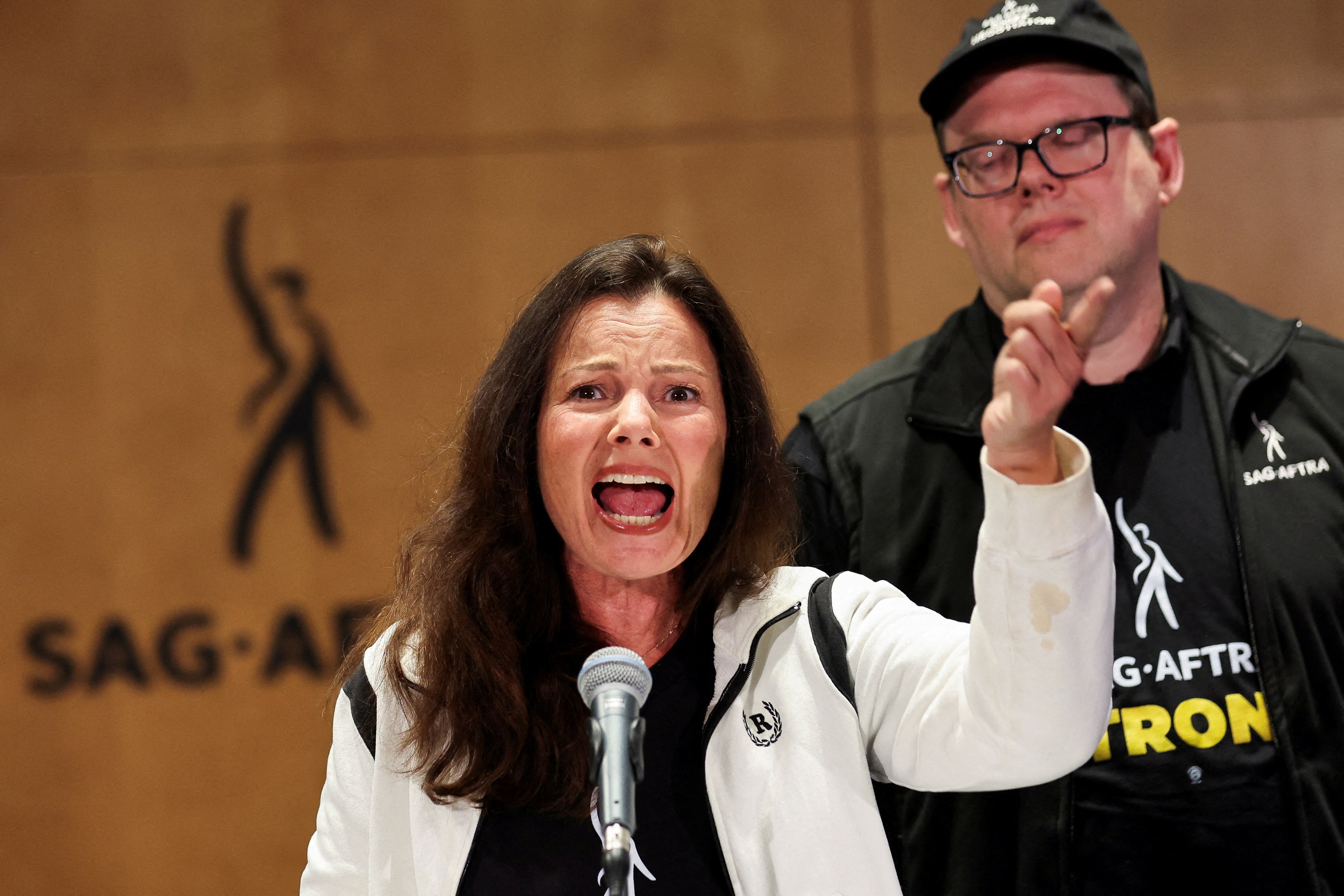 A woman speaks passionately into a microphone. She is holding up her right arm. A man stands behind her.