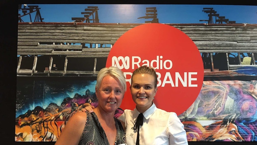 Blonde haired woman with brown haired woman standing in front of a sign for ABC Radio Brisbane