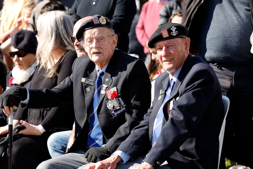 two men wearing suits and war medals sit on chairs in a crowd