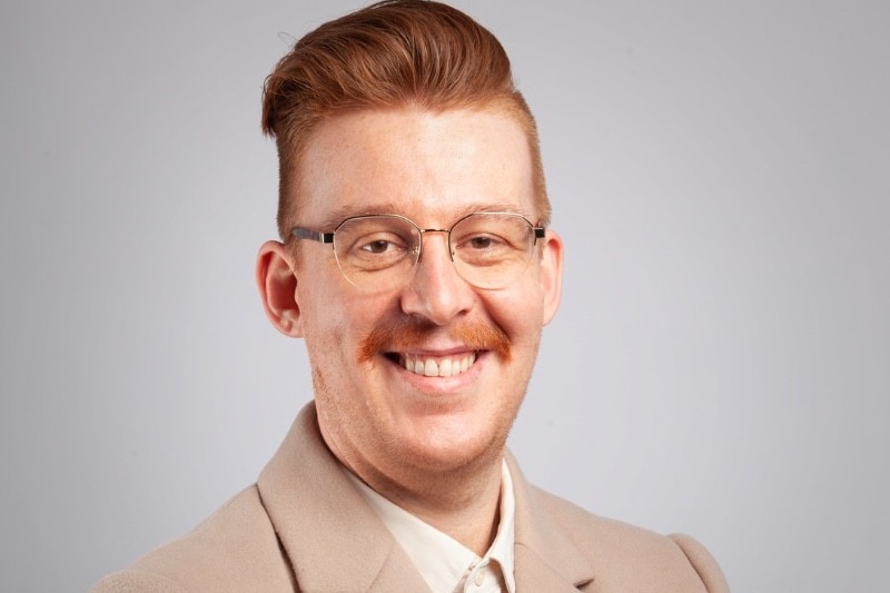 A headshot of man with red hair and mustache smiling, wears a beige suit, cream shirt, glasses.