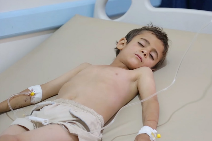 A young boy lies in a hospital bed with his eyes closed.