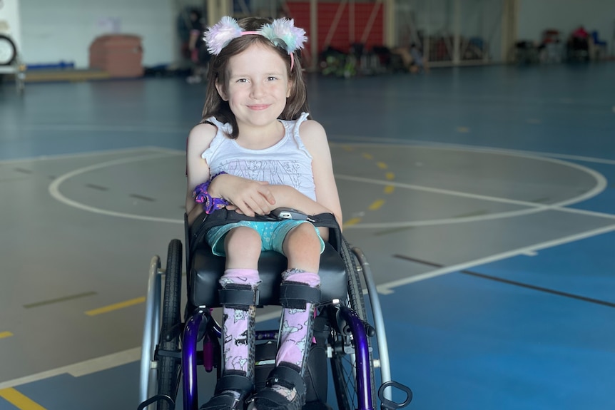 A young girl with a fluffy headband sits in her wheelchair on a basketball court