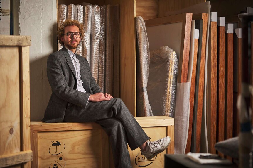 Gallery owner Michael Bugelli sits on a wooden box in a warehouse.