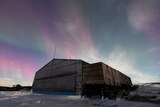 A pink and green aurora stretches over the sky above a shed in Antarctica. 