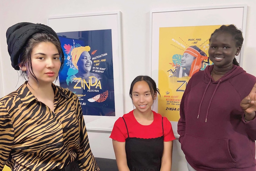 Three women stand in a room and look to the camera. They each smile, the woman on the right does a peace sign.