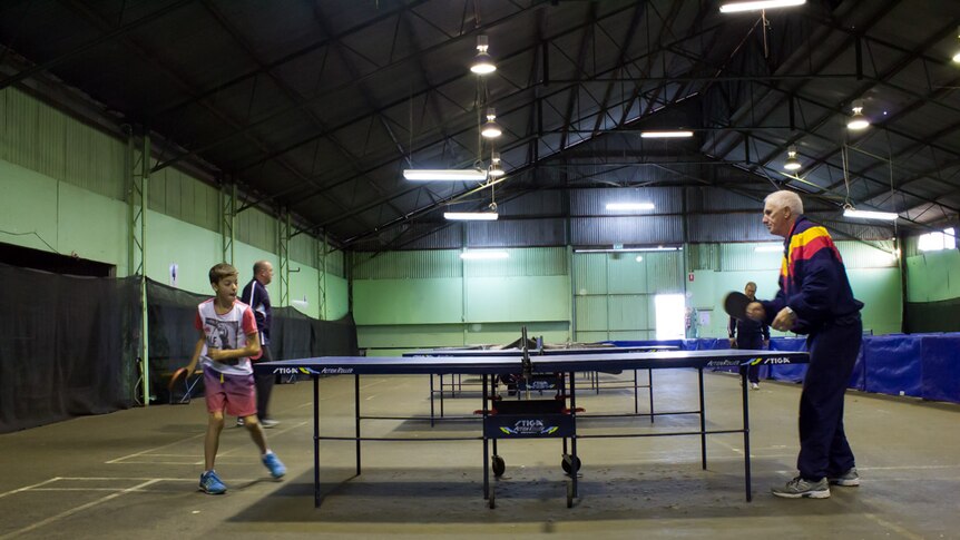 Players practice table tennis in a shed with a row of table tennis tables down the centre.