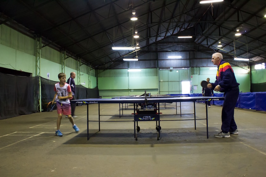 Players practice table tennis in a shed with a row of table tennis tables down the centre.