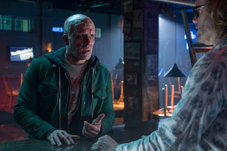 Colour still from 2018 film Deadpool2, Ryan Reynolds and T.J. Miller converse in a darkened and closed bar venue.