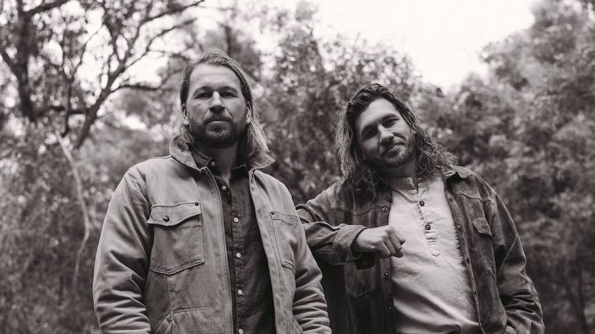 Black and white photo of Josh and Sam Teskey standing outside in a bush setting. They both wear jackets and look at the camera.