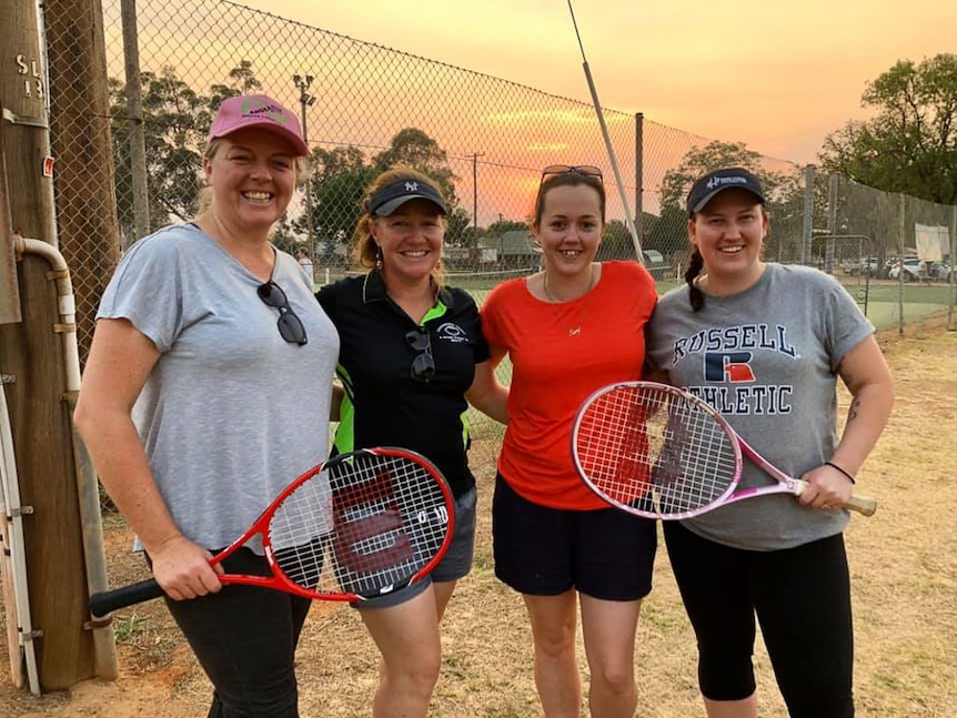 Four women holding tennis racquets in front of tennis court.