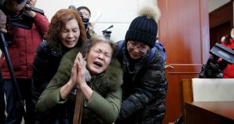 Wang Yulian is held up by two women as she holds her hands in a prayer position with photographers behind them.