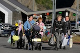 a group of people walk down a road holding bags and suitcases, one woman is pushing a pram