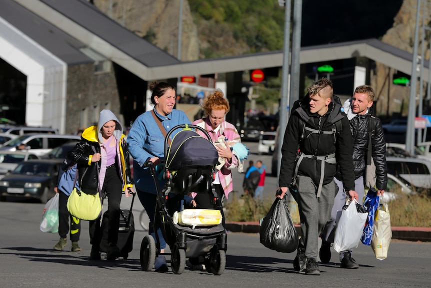 a group of people walk down a road holding bags and suitcases, one woman is pushing a pram