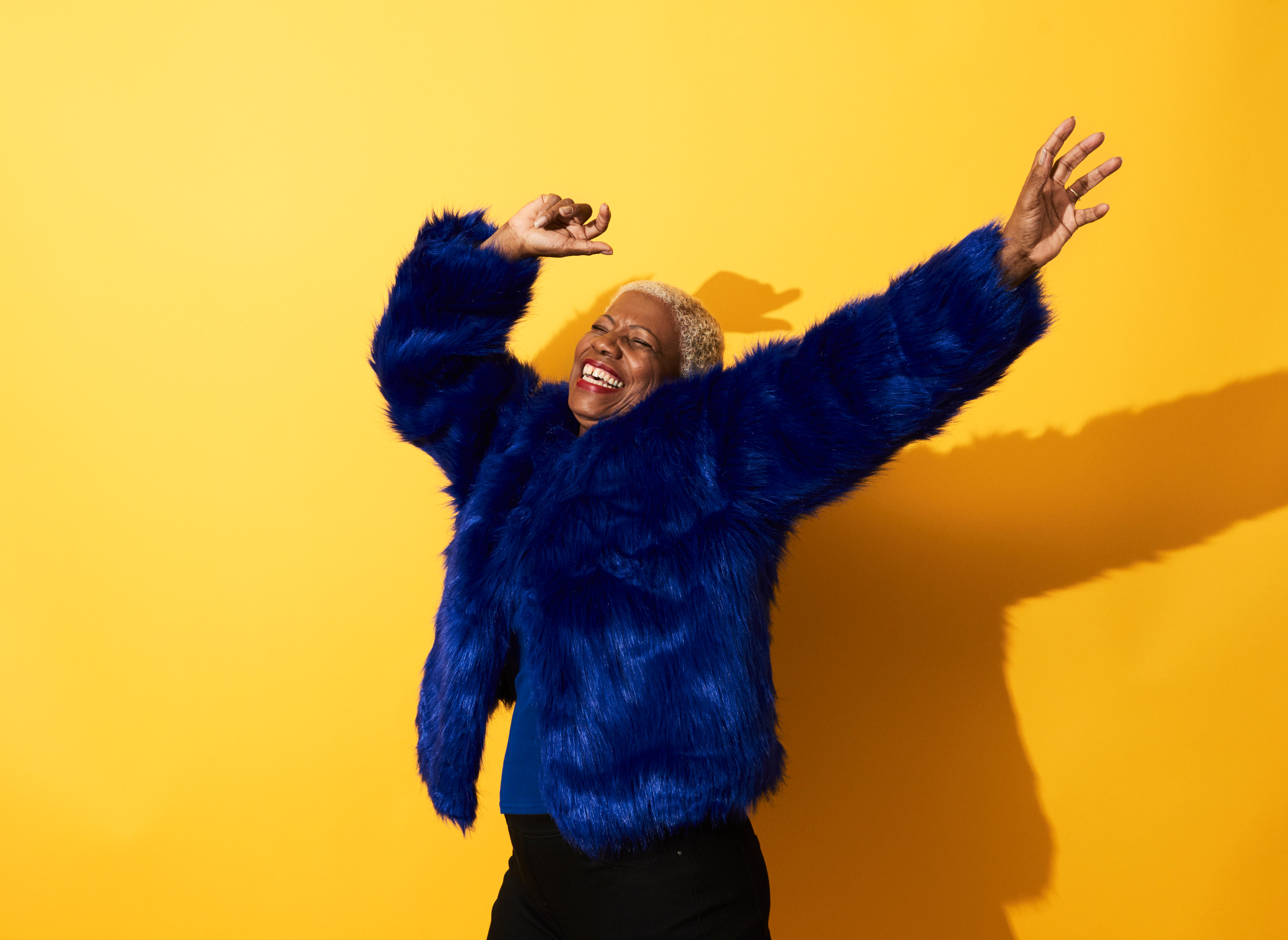 Black older woman in blue fur coat smiling with arms raised