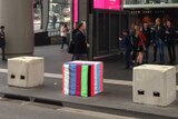 A series of large concrete blocks outside a busy railway station, one covered in stripy colourful material.