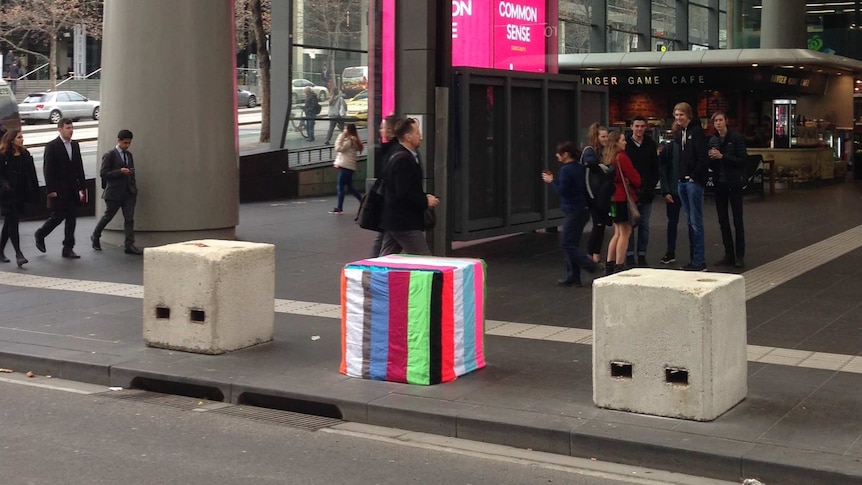A series of large concrete blocks outside a busy railway station, one covered in stripy colourful material.