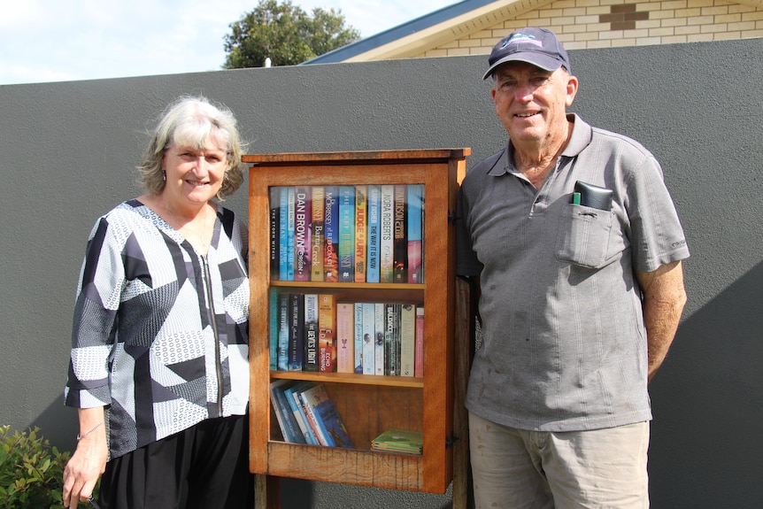 A man and a woman smile at the camera while standing next to a wooden cupboard filled with books