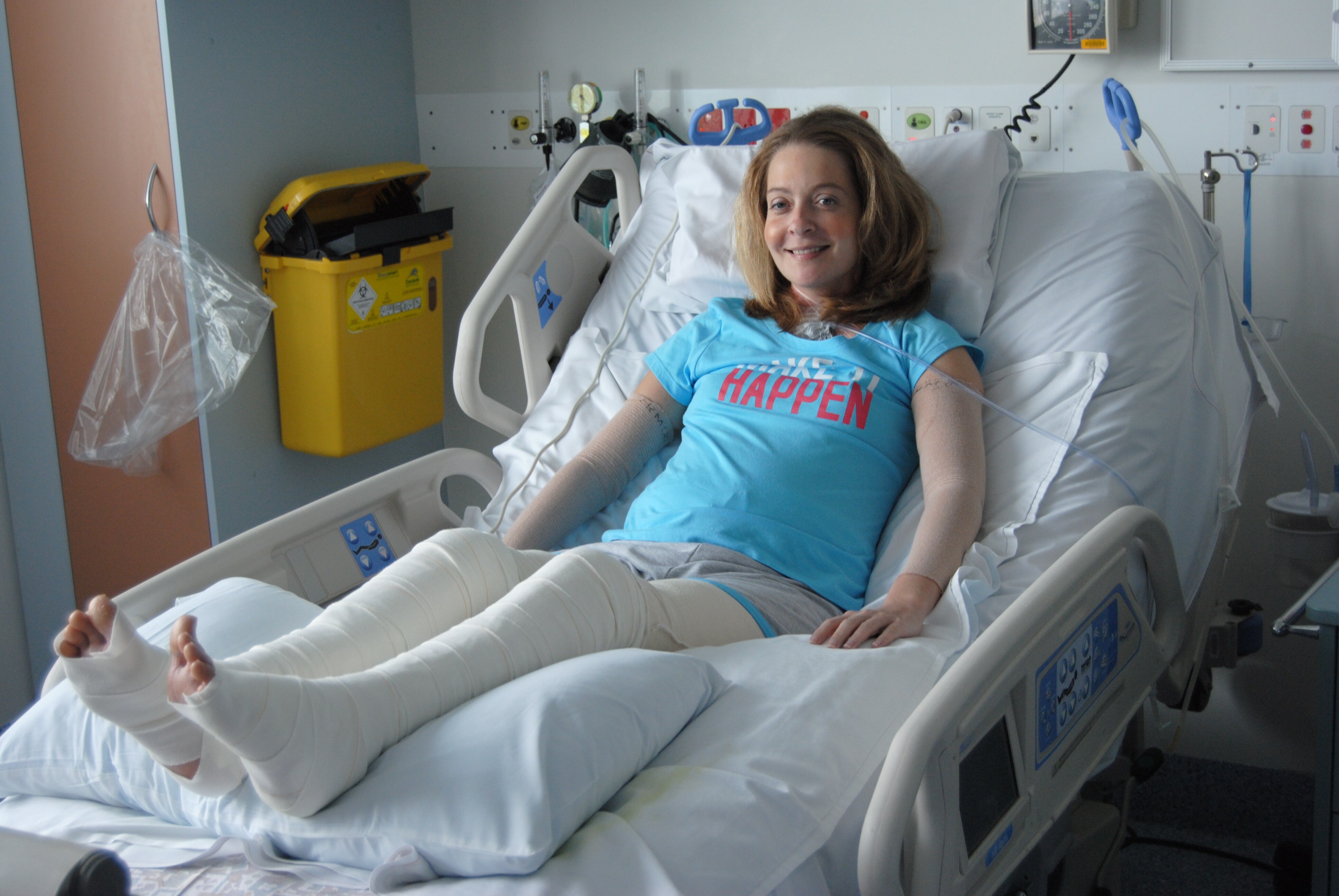 Una mujer sonriente en una cama de hospital. Tiene las piernas vendadas.
