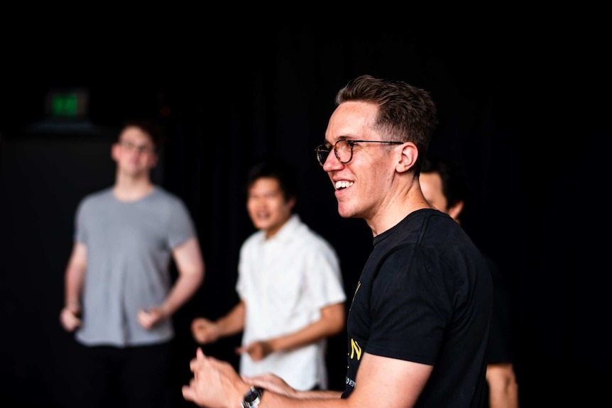 Man in black t-shirt, seen standing, side on, other people (blurry) in background