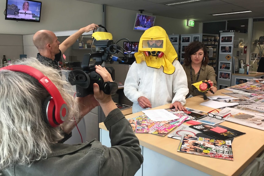 Paul Barry wearing yellow gas mask while being filmed in Media Watch office.