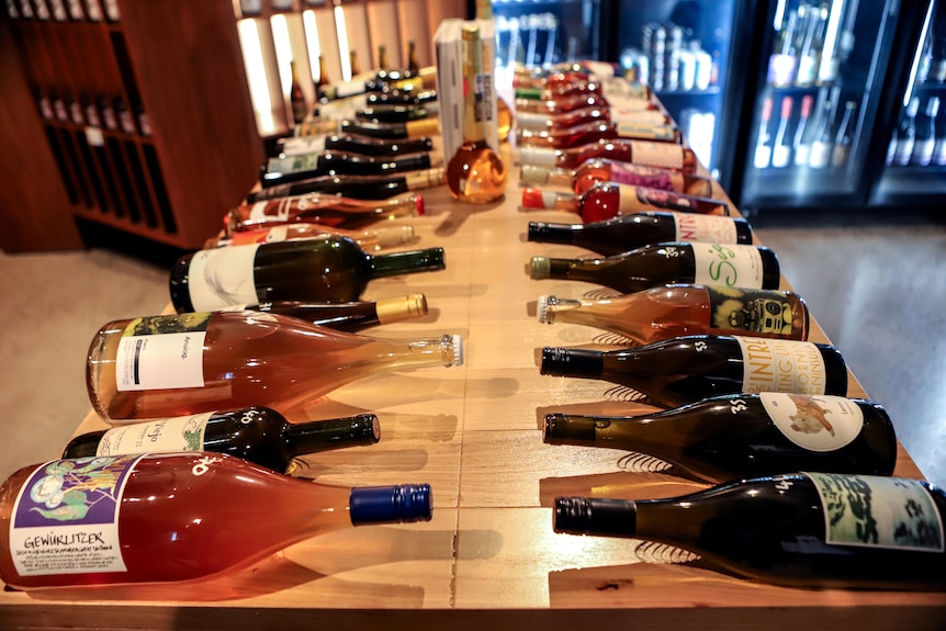 Two rows of brightly coloured wine bottles lined up on a wooden bench with an illuminated wine store fridge in background