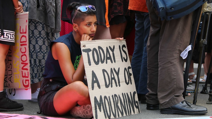A woman sits holding a hand-written sign saying "today is a day of mourning".