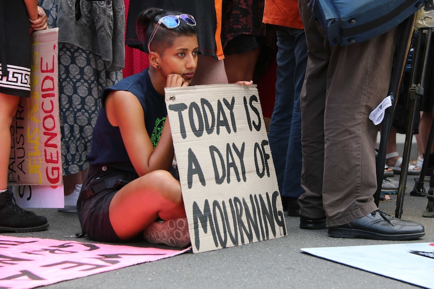 A woman sits holding a hand-written sign saying "today is a day of mourning".