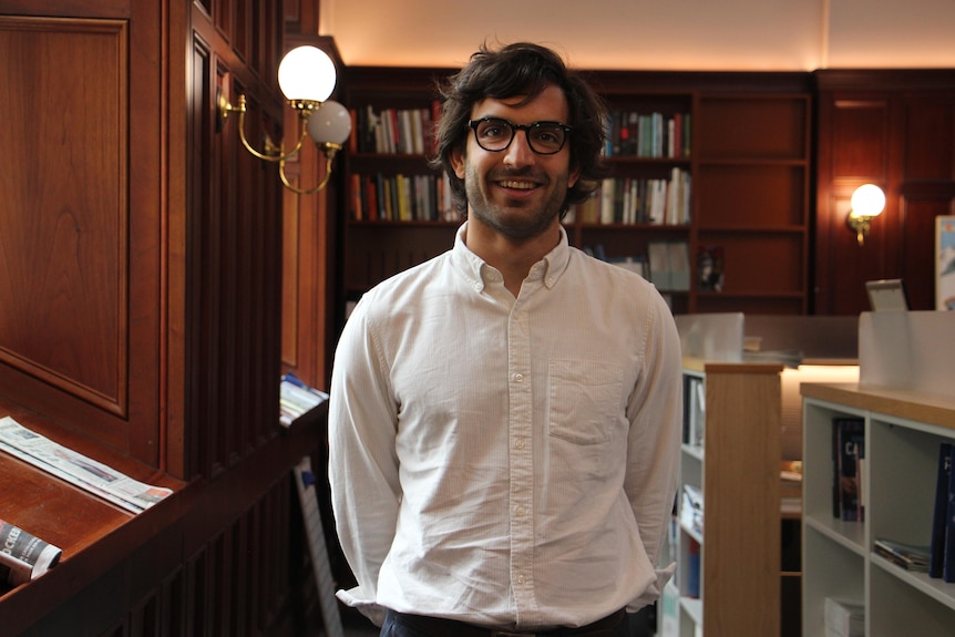 A man stands in an office. There are bookshelves and newspapers in the background.