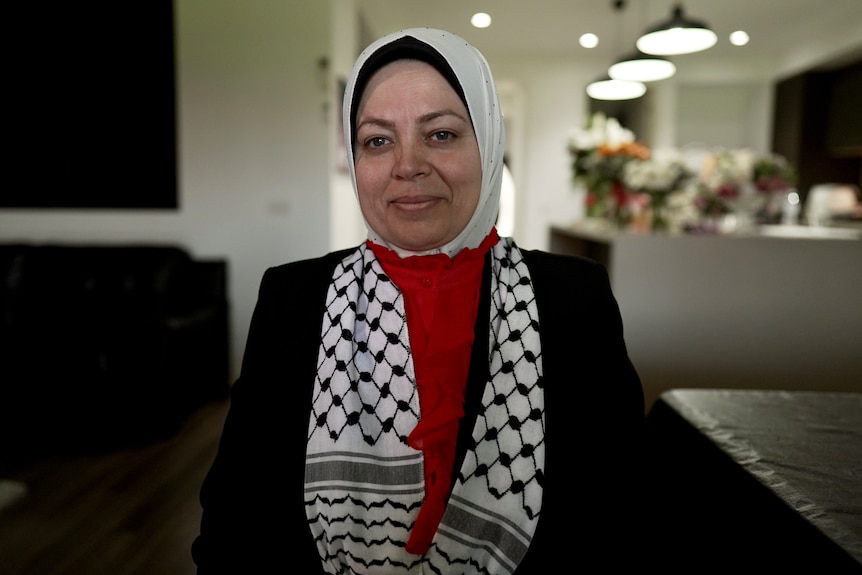 Muslim woman wearing a white hijab, red shirt and black jacket, sitting in her home.
