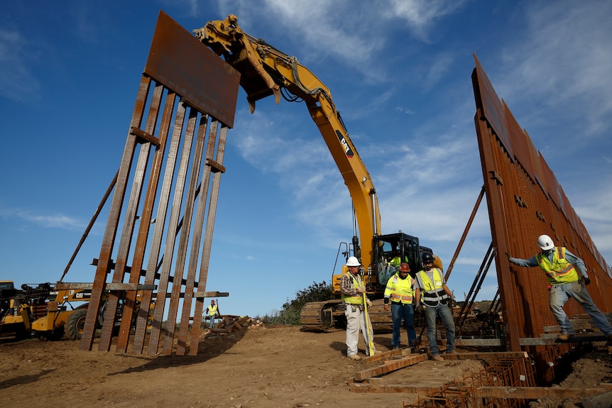 Four construction workers install large rusty sections of border wall with heavy machinery.